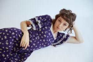 Stella Donnelly liegt auf dem Boden, ihr Kopf auf den aufgestützten Arm gelegt. Blaues Kleid mit Rüschen